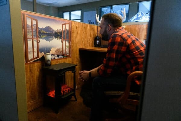 Mr. Mundt, wearing a red and black flannel shirt, sits in his cubicle, staring at a picture of a window that opens onto the scene of a lake in the mountains.