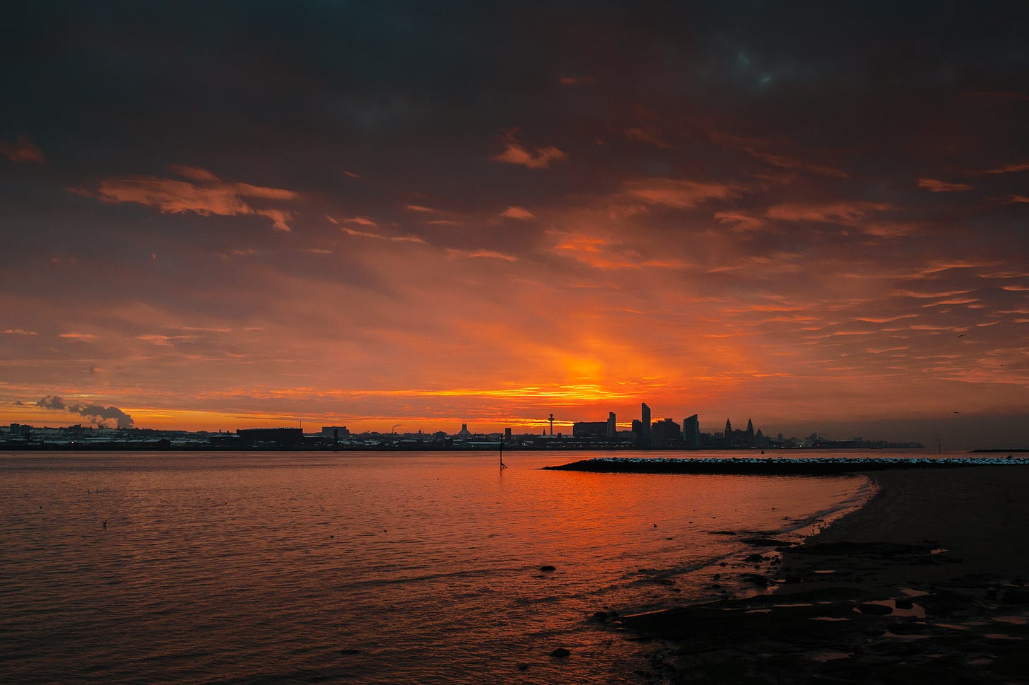 As the sunrises the sky is filled with deep red colours and clouds. Across the River Mersey there are buildings and homes covered with snow.
