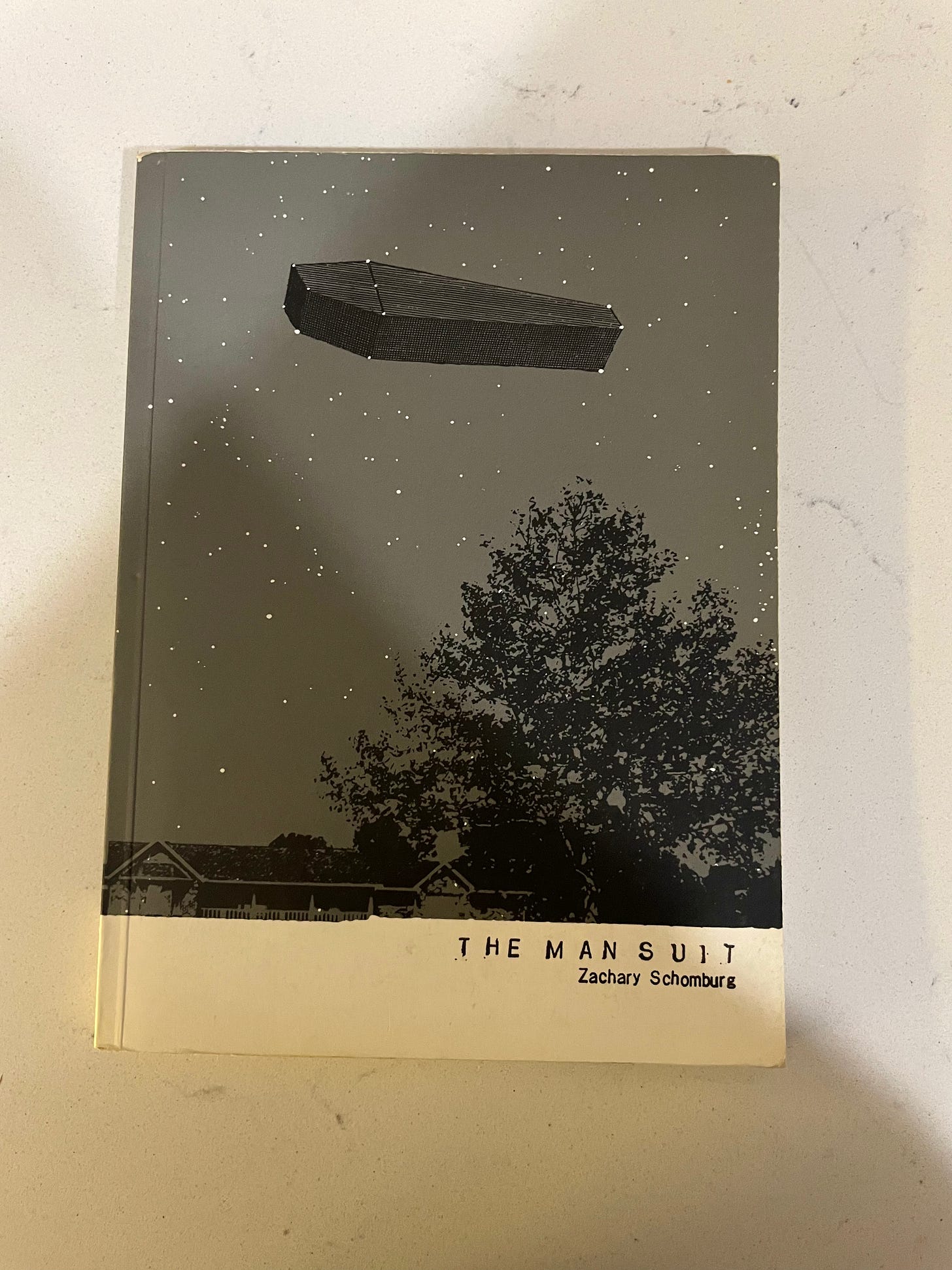 'The Man Suit' by Zachary Schomburg. Cover is a gray sky with white stars and a black tree and a black ranch-style house with a gray-on-darker-gray coffin floating in the sky