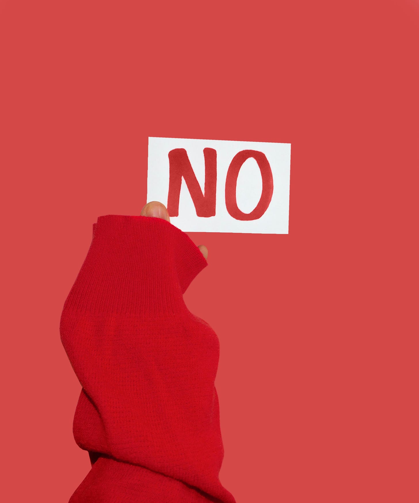 A hand in a read sweater, holds up a white card with the word “no” in red ink, against a red background. 