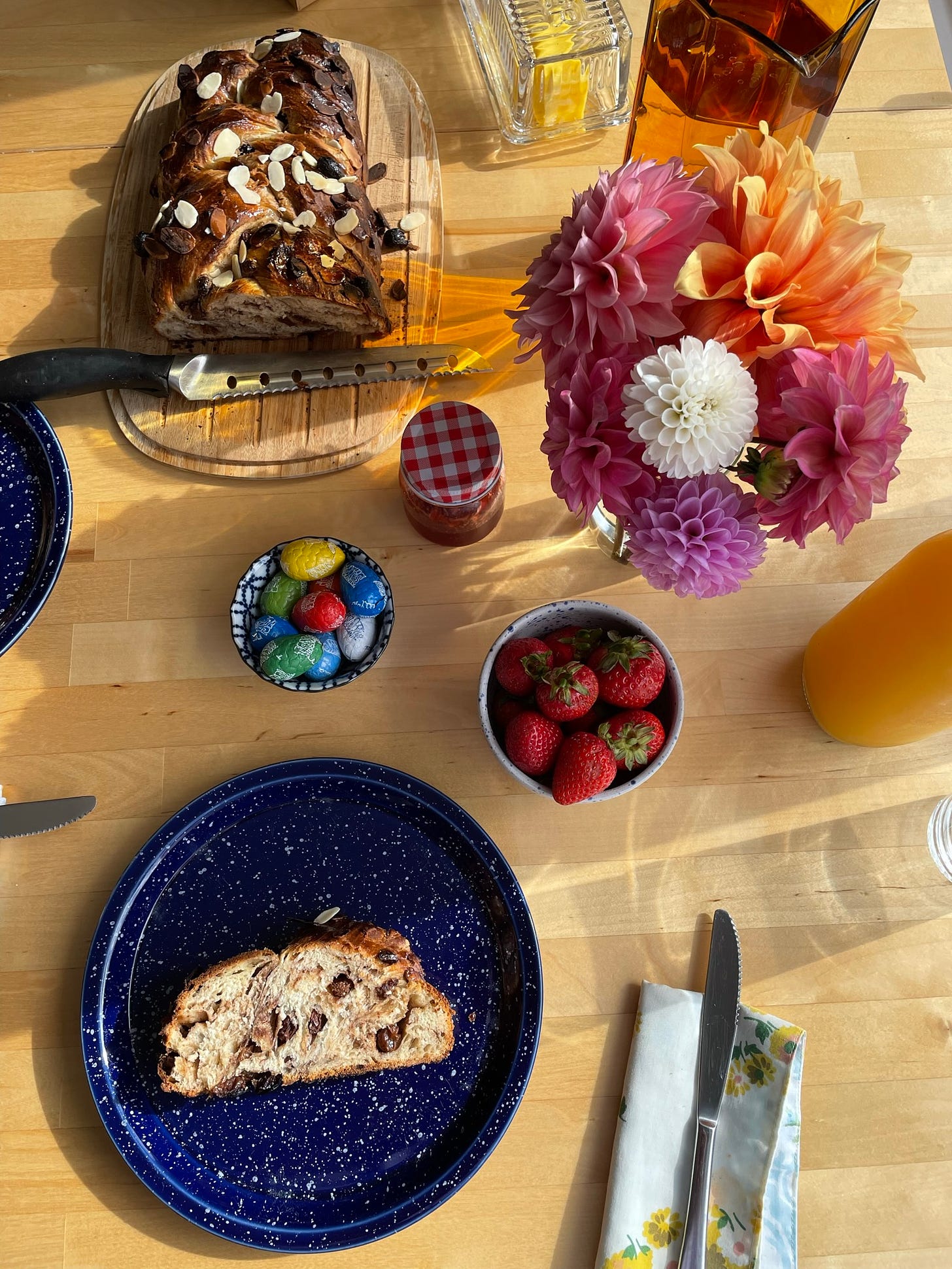 Table setting in morning sun with a braided fruit loaf, small vase of pink, orange and white dahlias, easter eggs and strawberries