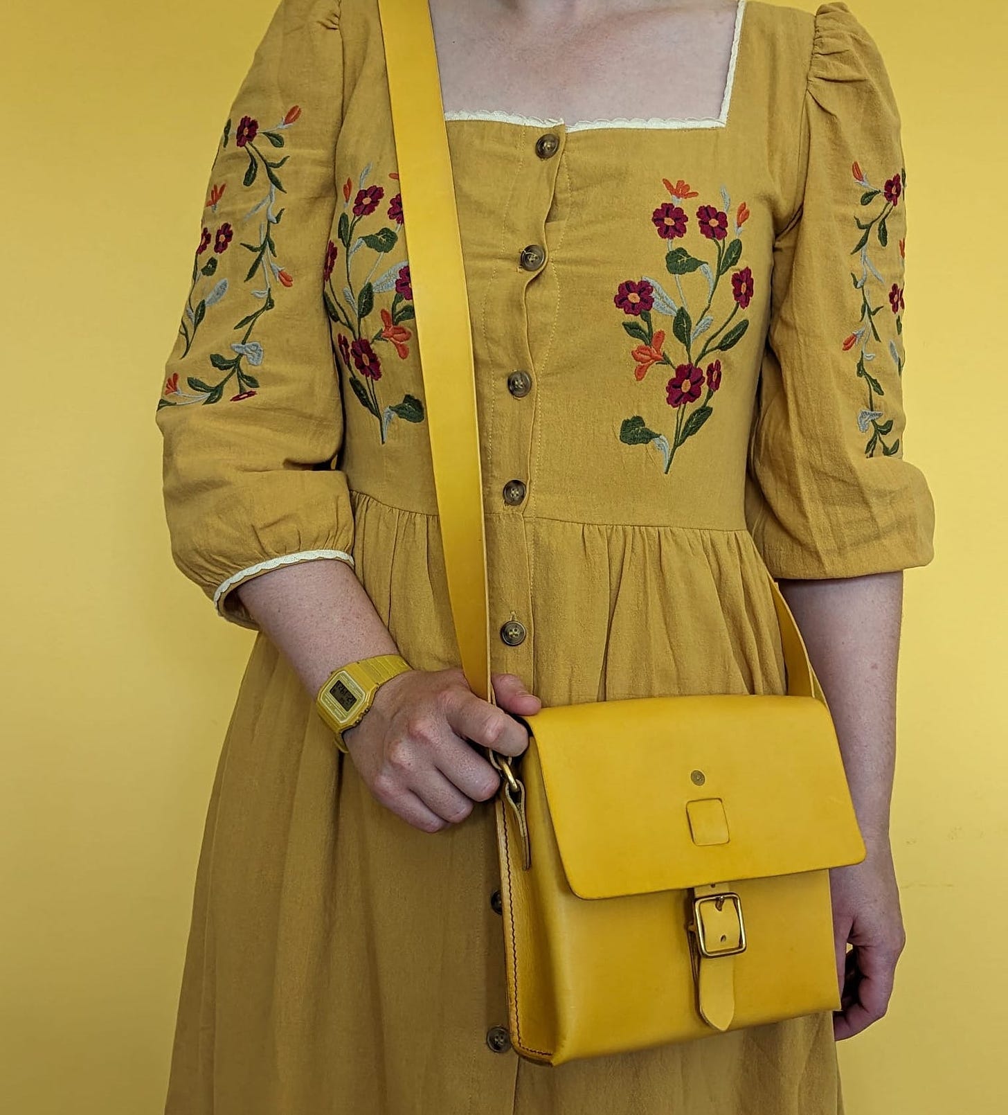 A cropped photo of Libby wearing a yellow dress embroidered with colourful flowers, holding a yellow cross-body bag and with a matching yellow watch. She is standing against a yellow wall. 