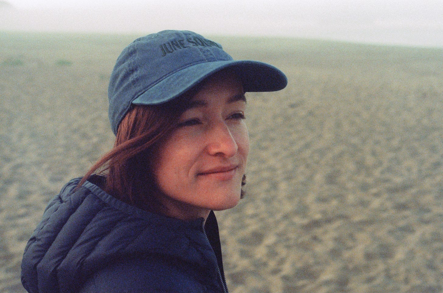 A portrait of Sarah standing near the ocean. She wears a blue jacket and baseball cap, and smiles gently while looking away from the camera.