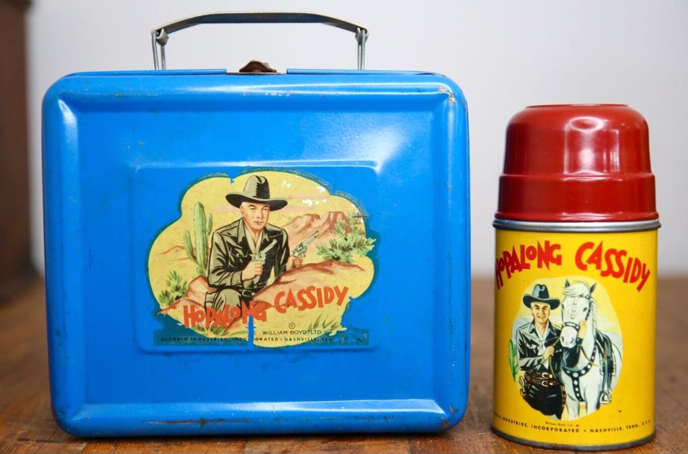Photo of a vintage blue tin lunchbox with a Hopalong Cassidy decal on the side. There’s also a Hopalong Cassidy branded Thermos with a red plastic lid. Photo of a vintage blue tin lunchbox with a Hopalong Cassidy decal on the side. There’s also a Hopalong Cassidy branded Thermos with a red plastic lid.