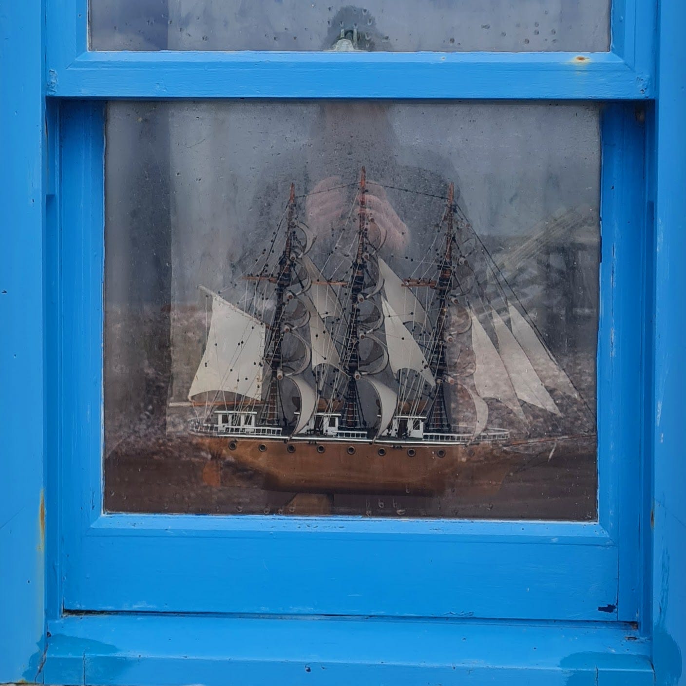 Photo of a blue painted window frame with a model ship inside, and a reflection of me in the window glass.