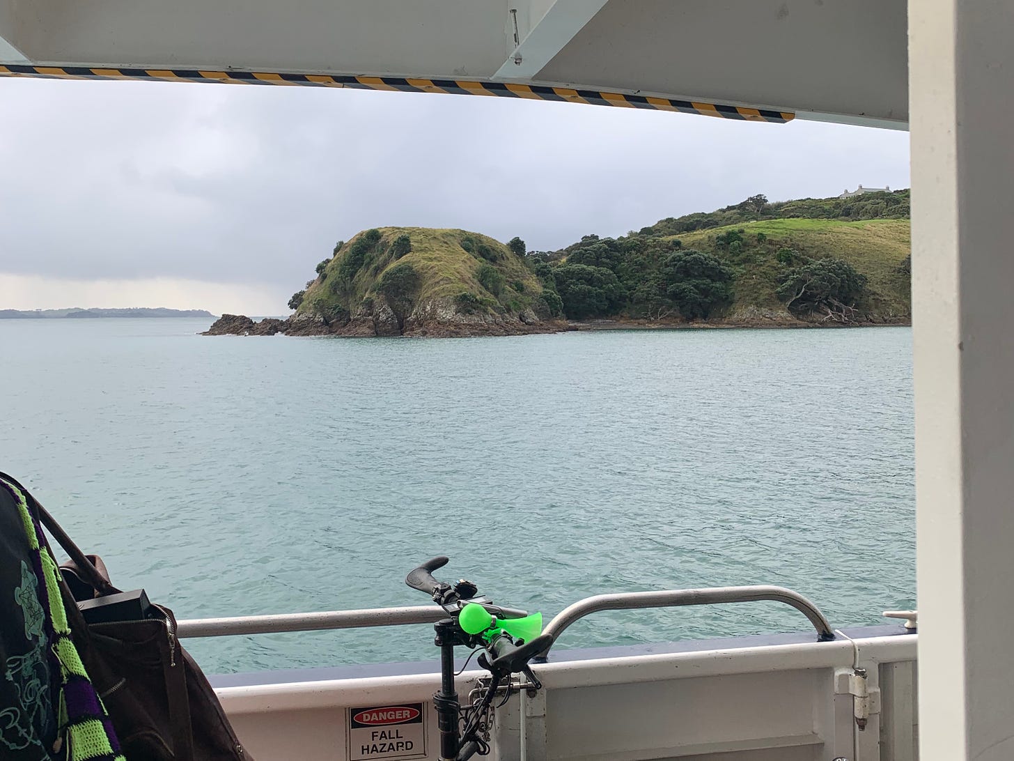 Matiatia headland on Waiheke Island, as framed through the upper and lower decks of the passenger ferry as it enters the harbour. In the foreground are the handlebars of a bike with a little green horn. A small sign on the ferry reads "fall hazard" and on the upper frame of the view is a strip of yellow and black danger tape. It is an overcast day but the headland glows a moss green. The sea is a dull teal and the sky is a grey cloud with a small amber band of clearer sky on the horizon above Rakino, another island in the Hauraki Gulf