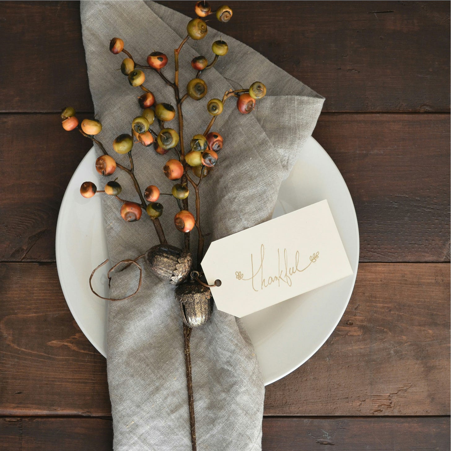 A table setting with a white plate, gray napkin, sprig of acorns, and a paper note that reads "thankful."