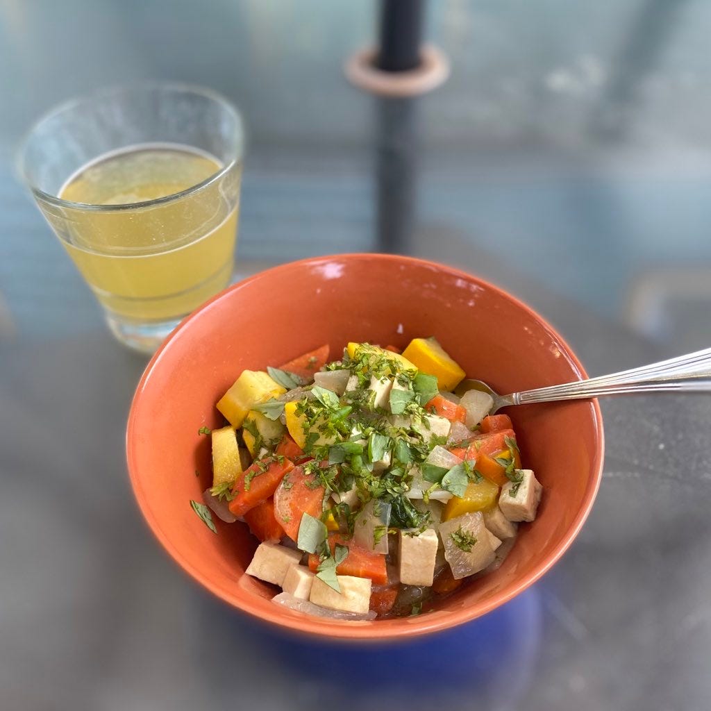 An orange bowl full of green curry with tofu, carrots, and yellow zucchini, over jasmine rice. On top is roughly chopped cilantro, and a fork sticks out of the bowl at the edge. Above and to the left of the bowl is a glass of pale, hazy beer.