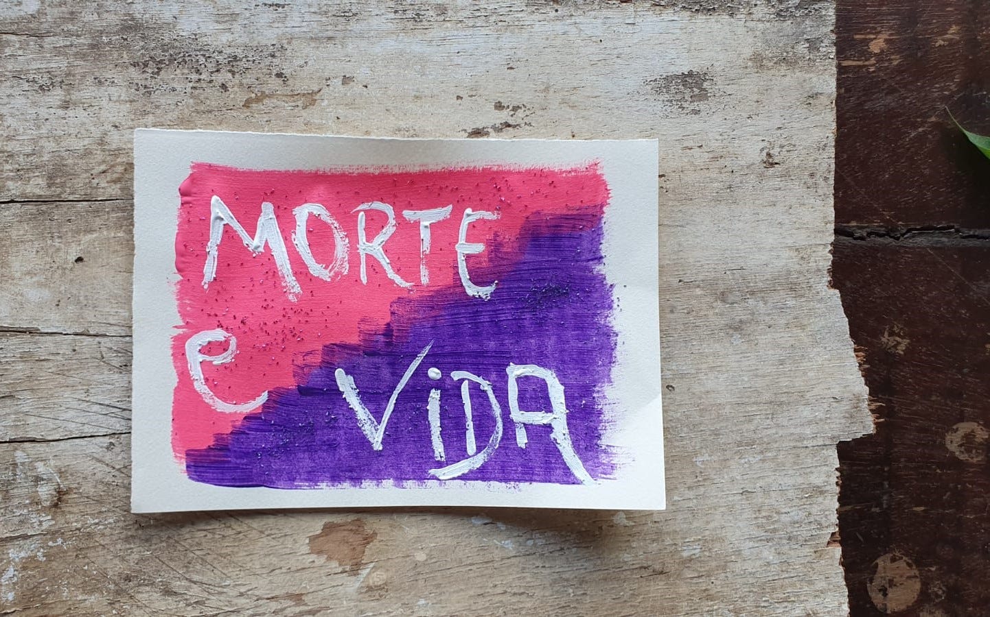 Fotografia de um papel pintado de rosa e roxo sobre um fundo de madeira com o escrito "Morte e Vida"