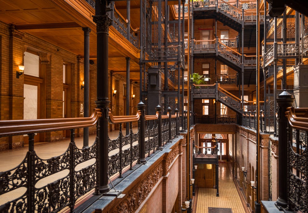 Interior photo of the Bradbury Building. It is wood and brick, with metal railings. Classy in style, lit by lanterns. Multiple floors are visible, as well as the ground floor below. There are multiple narrow halls, stairways, and the like leading to a complicated but interesting internal design aesthetic