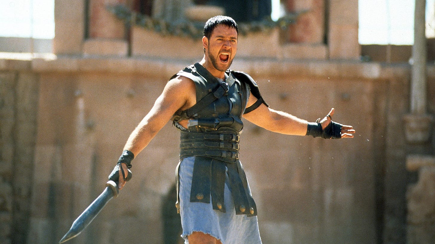 Still from Gladiator. Russell Crowe as Maximus holds his arms wide to the crowd, calling out "Are you not entertained?" without the aide of a public address system.