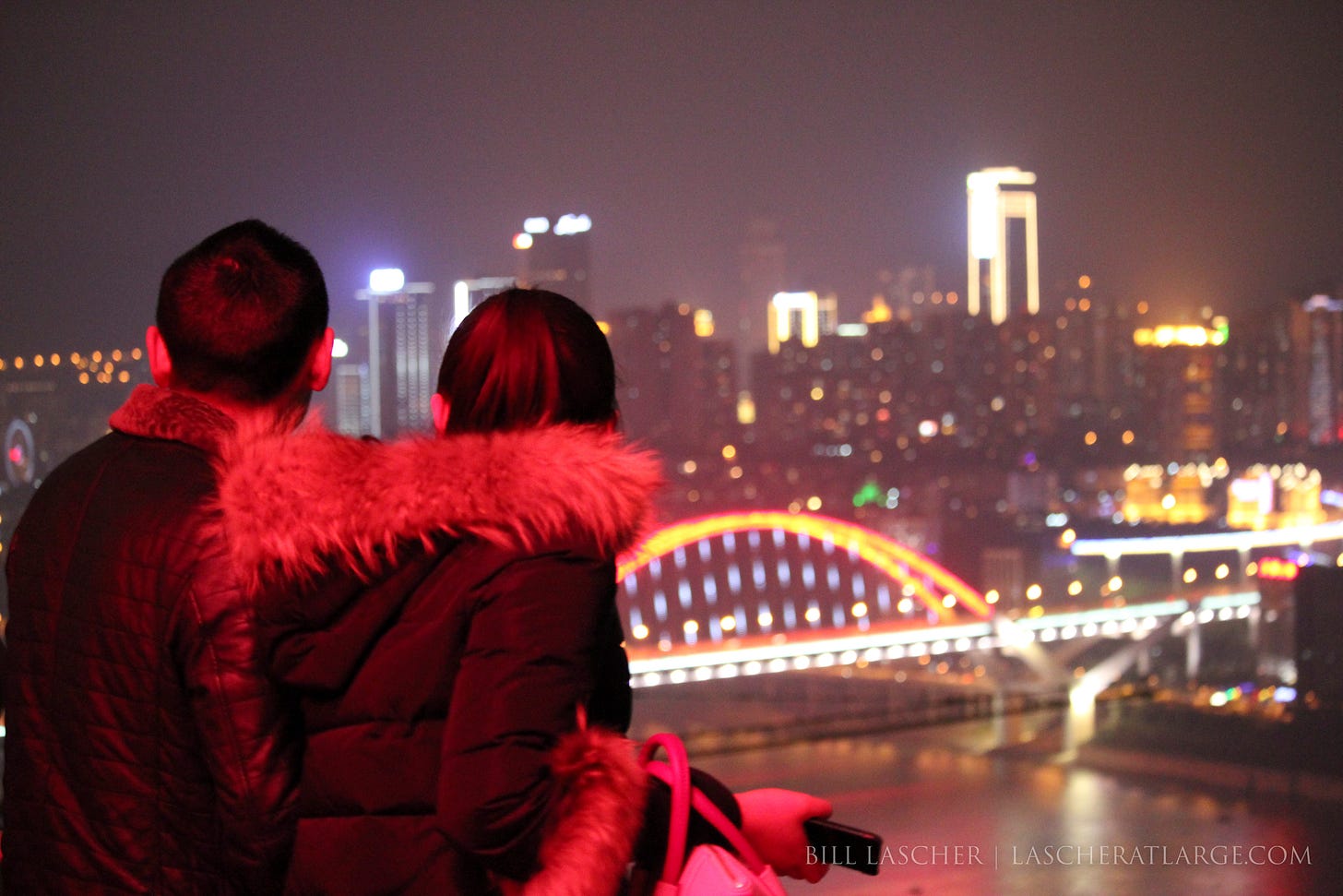A couple with their backs to the camera look over a brightly-lit urban skyline and an arc-shaped bridge illuminated with glowing lights crossing a river in the lower third of the photo. It is nighttime and many of the buildings have lines of bright lights around their edges. The couple's fur lined coats have a red tint cast by lights behind the photographer.