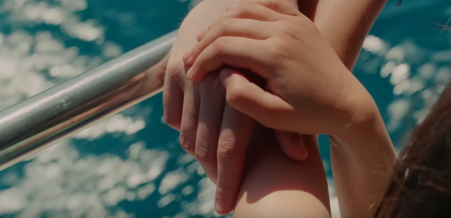 A larger adult hand holds a smaller hand, that of a child. They rest against a metal railing. In the background, you can see sun dappling across blue water.