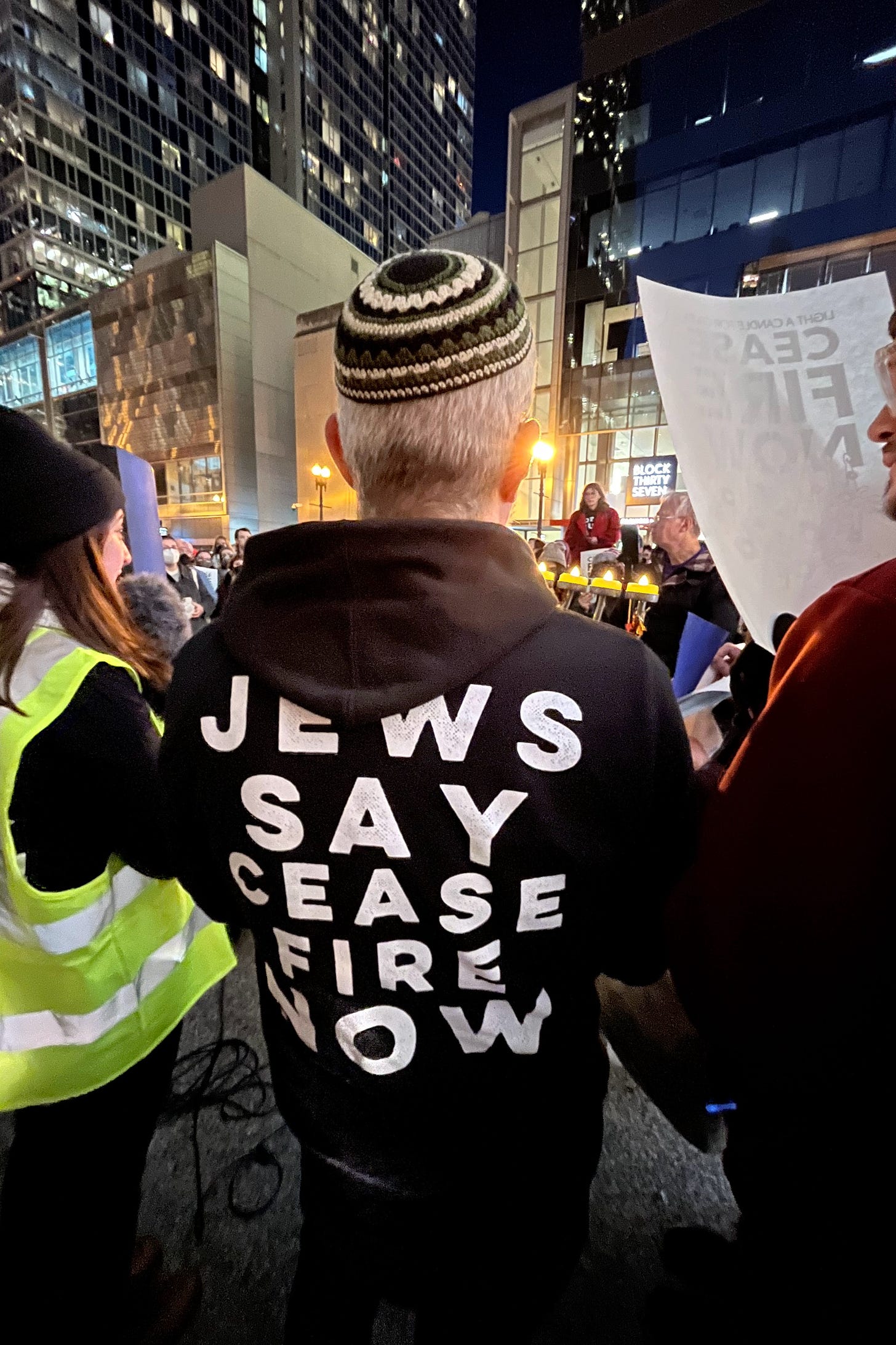 A rabbi facing away from the camera holds a menorah. The back of his sweatshirt says "Jews say ceasefire now"