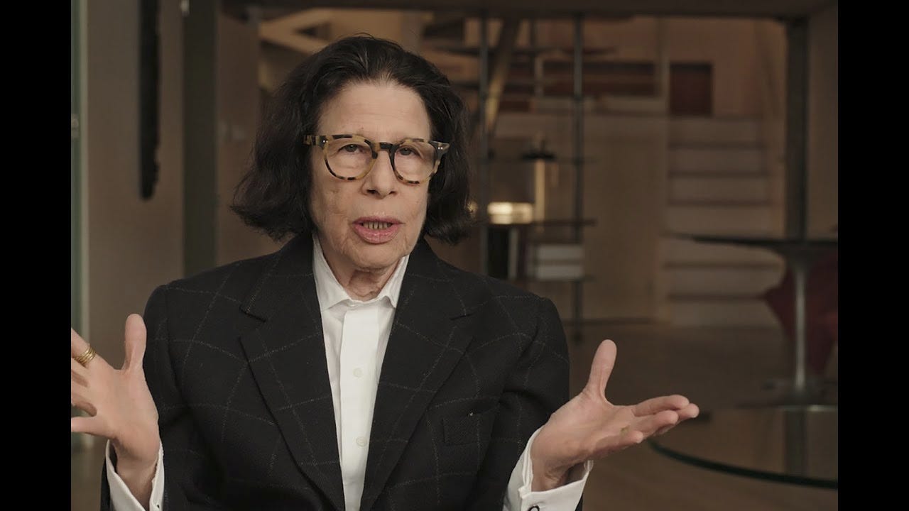 A screencap of Fran Lebowitz, a white woman with jaw-length black hair, tortoiseshell glasses, in a black blazer and white shirt, holding her hands out in a shrug of dismay. 