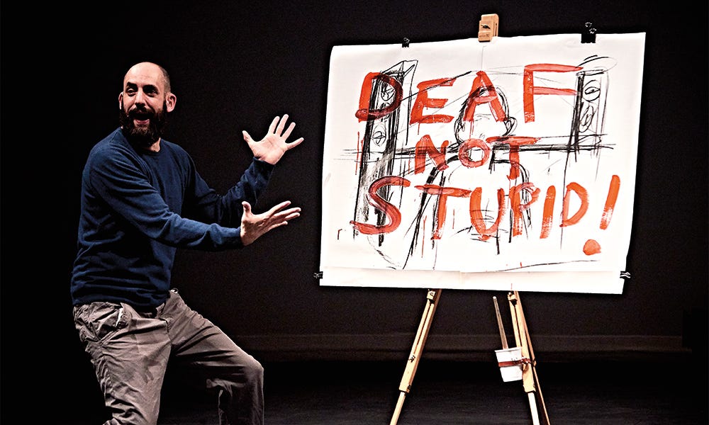 A white man on stage is presenting a big reveal on an easel. The canvas has a line drawing of a person and on top, it says DEAF NOT STUPID!