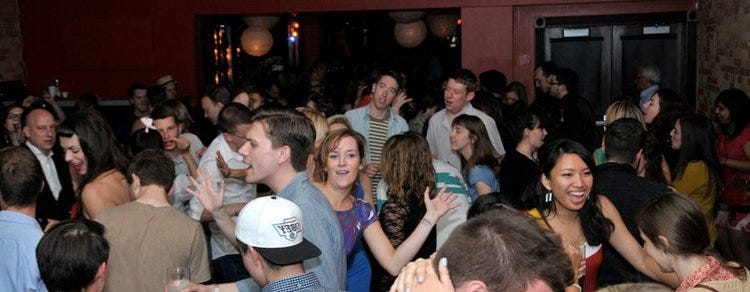 Dancefloor full of white people. Justine, the only racialized person, is smiling a big smile while dancing with her friends.