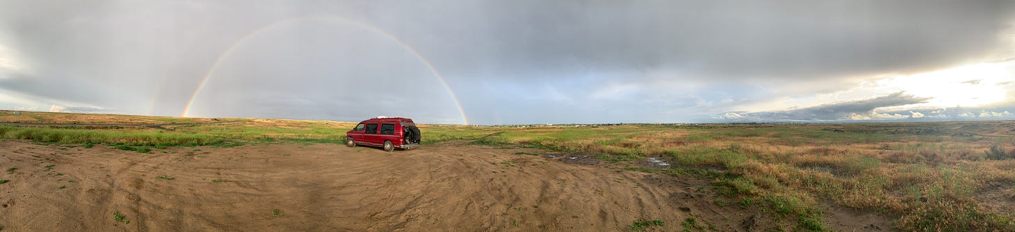 A panoramic photograph of a field with a ruby-colored van in the center underneath a double rainbow arc