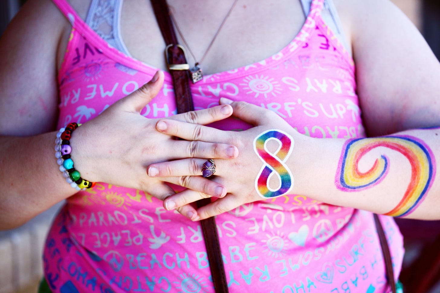 A fair skin person, close up of crossed arms on their chest sporting a rainbow swirl on on arm and a rainbow infinity on their hand. The person is wearing a pink top.