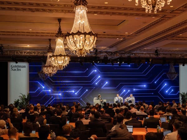 A ballroom of people watch as three people talk on a stage.