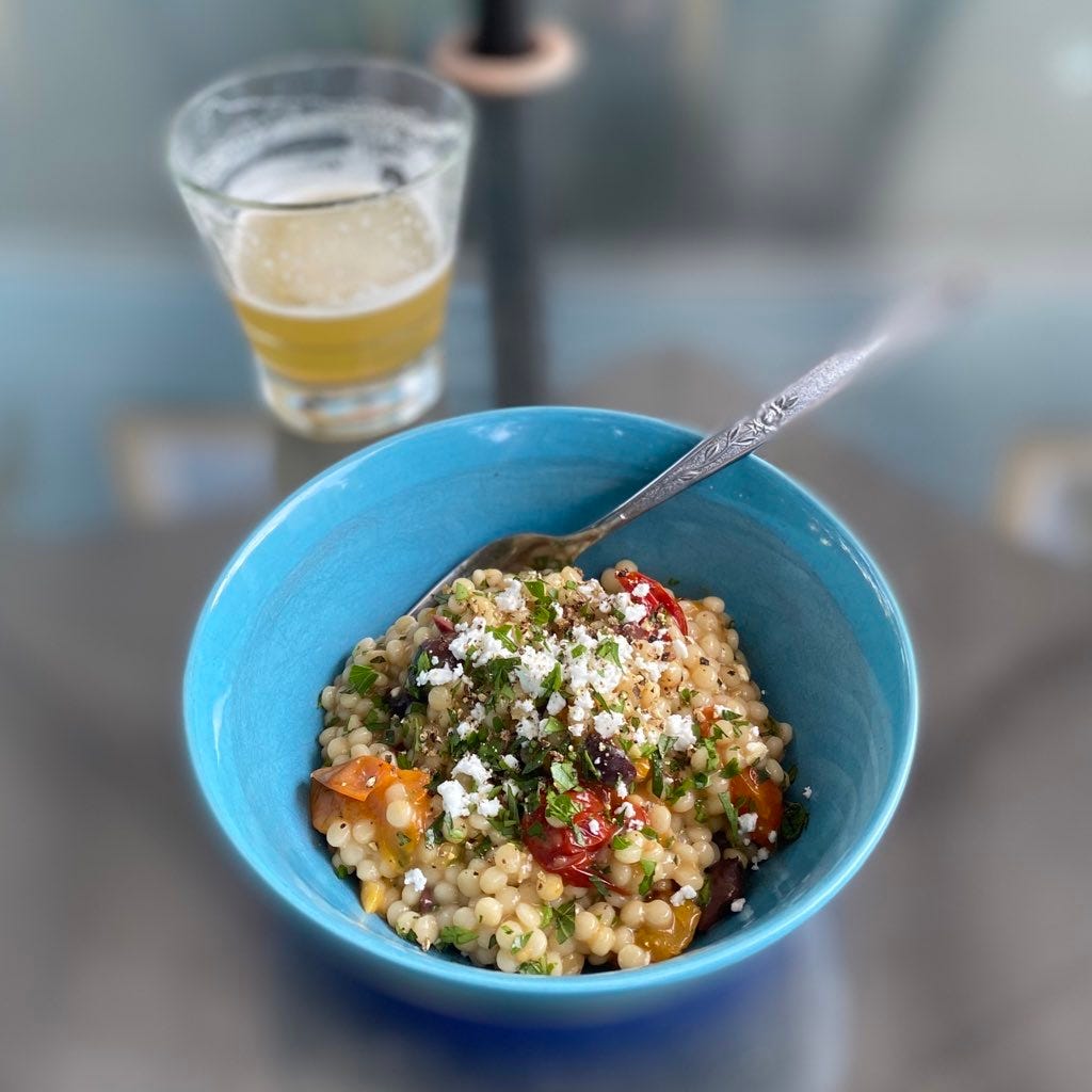 A blue bowl of the pearl couscous described above, with roasted multicoloured tomatoes, feta, and kalamata olives. A partially full glass of beer sits in the background.