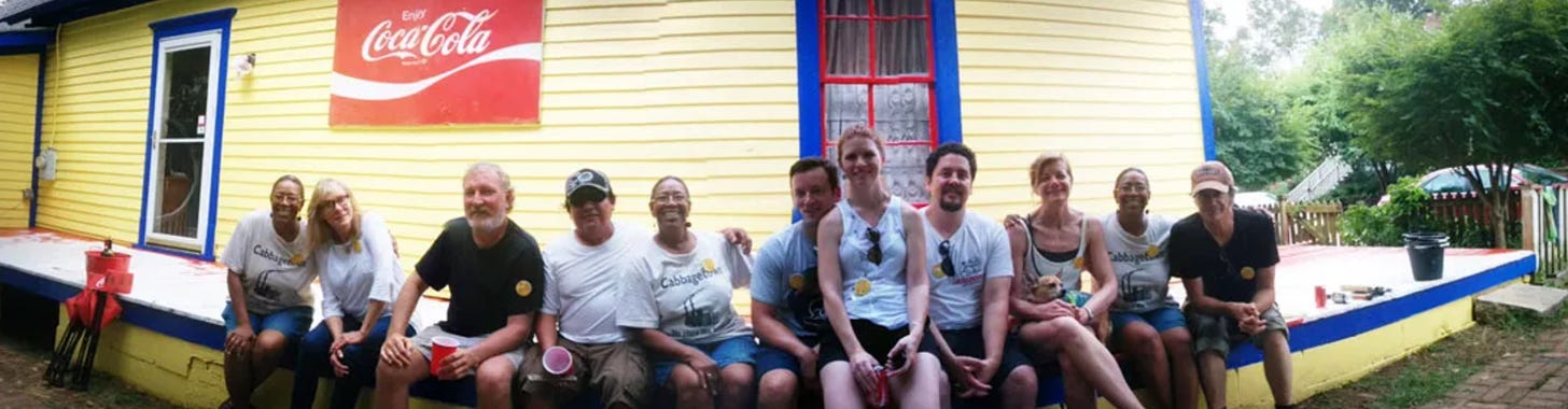 The volunteer painting crew on the porch of the "Coke House," yellow with blue and red trim. Owner Bertha can be seen on either side of the line and in the center.