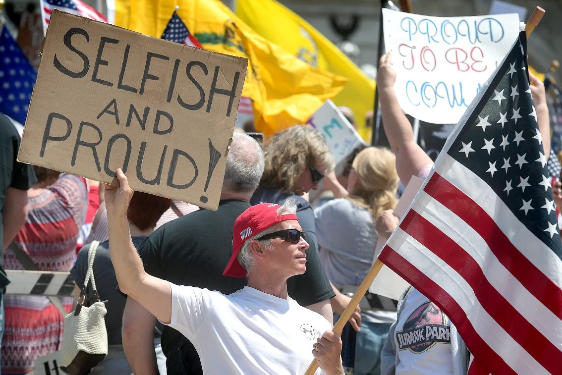 A white man brandishing an American flag and a sign reading "SELFISH AND PROUD" at a pro-COVID protest.