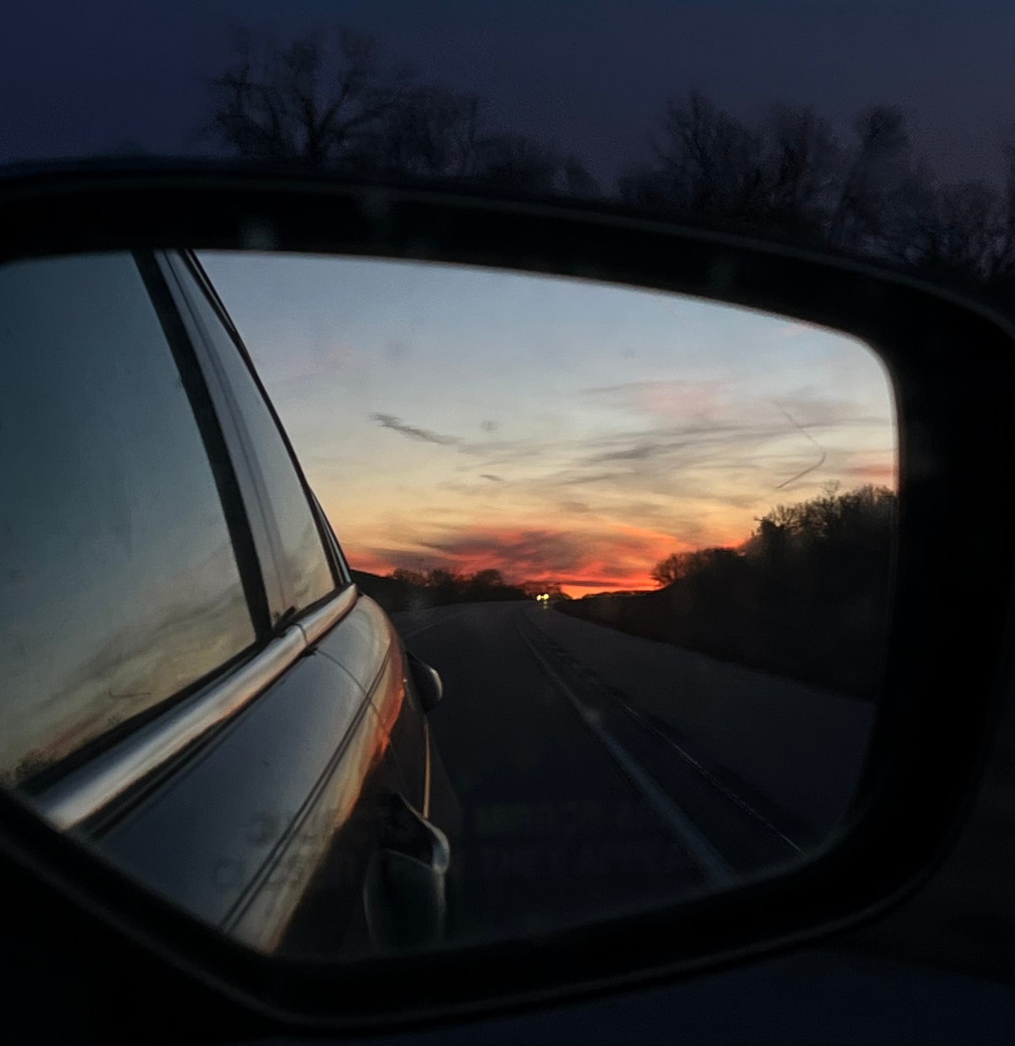 In a side mirror, the edge of the car is visible, and beyond is the road and sunset. Near the horizon, it's bright orange with clouds in front.
