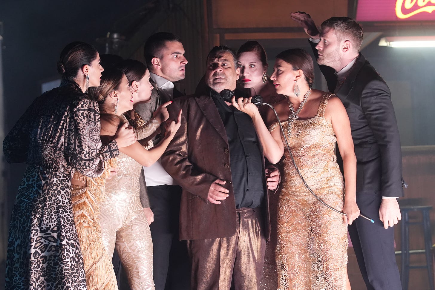 Scene from The Divine Comedy. A group of people in opulent evening wear crowd around a man in a bronze suit,