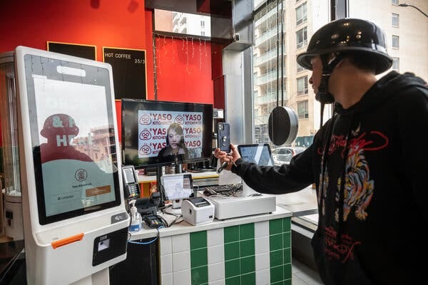 A man wearing a biking helmet adjusts a device attached to a monitor. On the monitor is a woman with dark hair wearing a headset in front of a backdrop that reads Yaso Kitchen. 