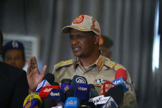 A man holds up his hand while speaking into several microphones placed in front of him.