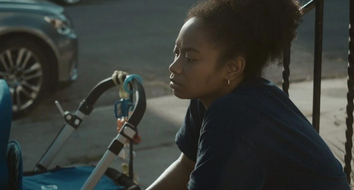 Still from a film. A Black woman sits with her elbows on her knees. She is wearing a black t-shirt and her hair is up in a bun. Her eyes are downcast. She is sitting on a stoop with a wrought iron railing behind her. A baby stroller is in the background.