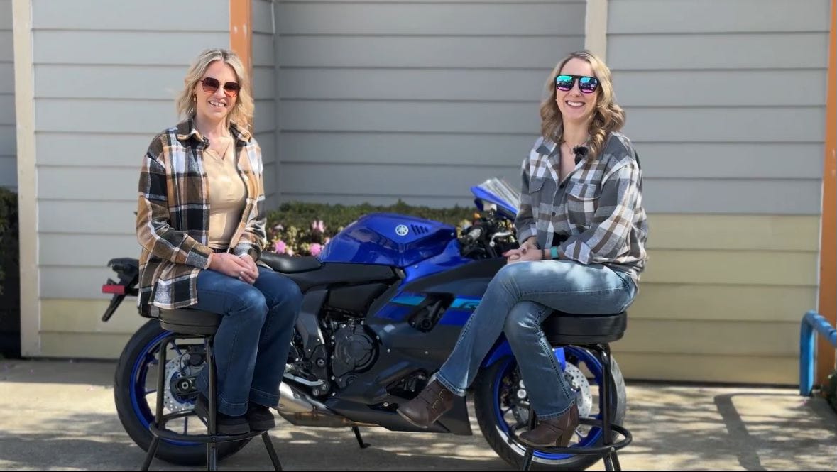 Two women sit in front of a blue Yamaha motorcycle