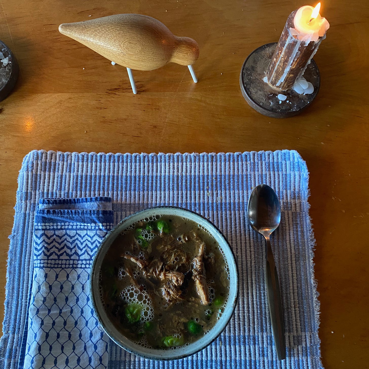 A bowl of steaming soup on a placemat, next to a lit candle and a small wooden sand piper figurine. 