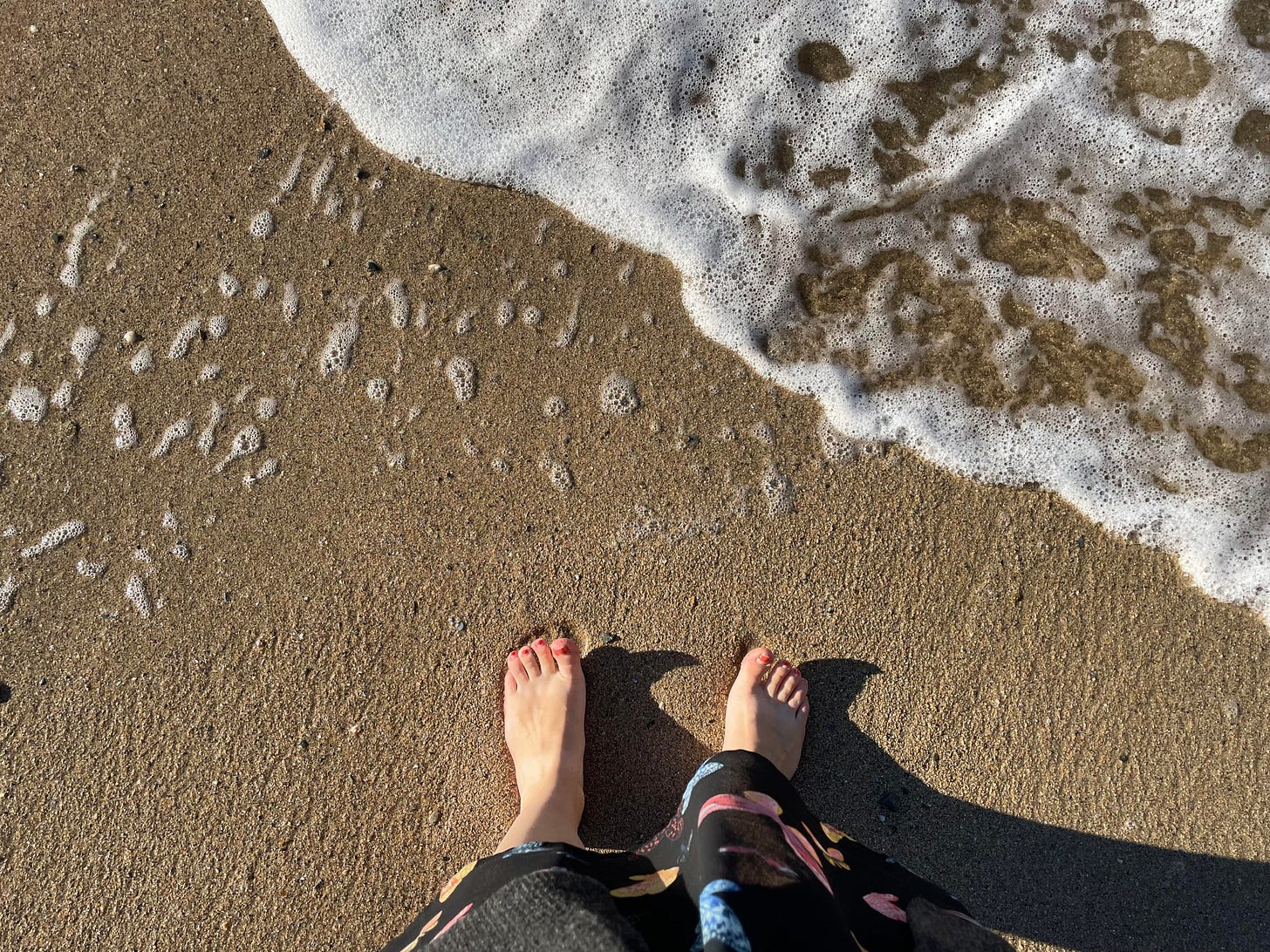 A person standing on a beach seen from above, with bare feet standing in the sand next to some foam from the sea water