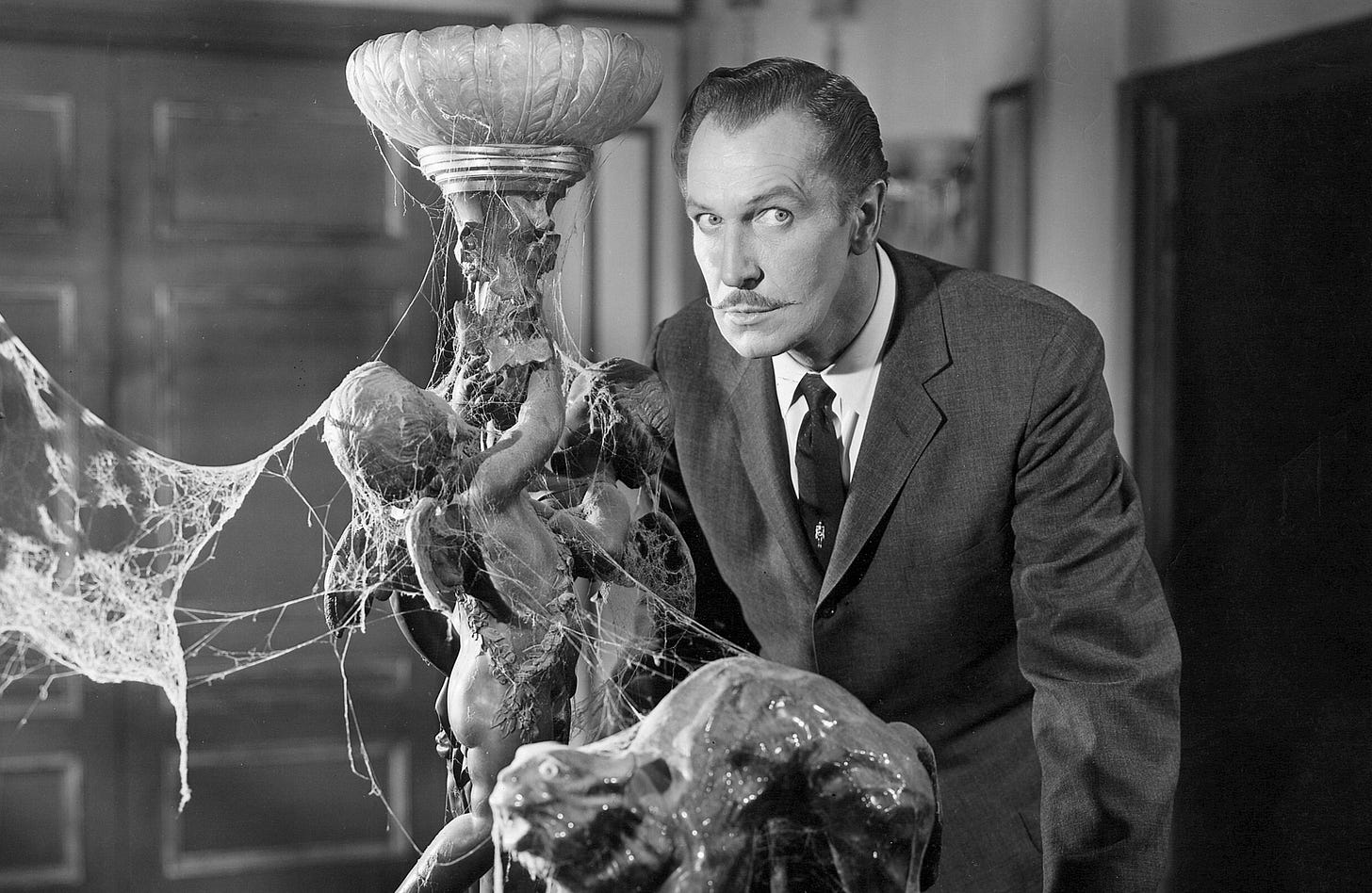 Shot of Vincent Price from House on Haunted Hill. He's looking directly into the camera (and into your soul) while standing behind a decorative lamp covered in cobwebs.