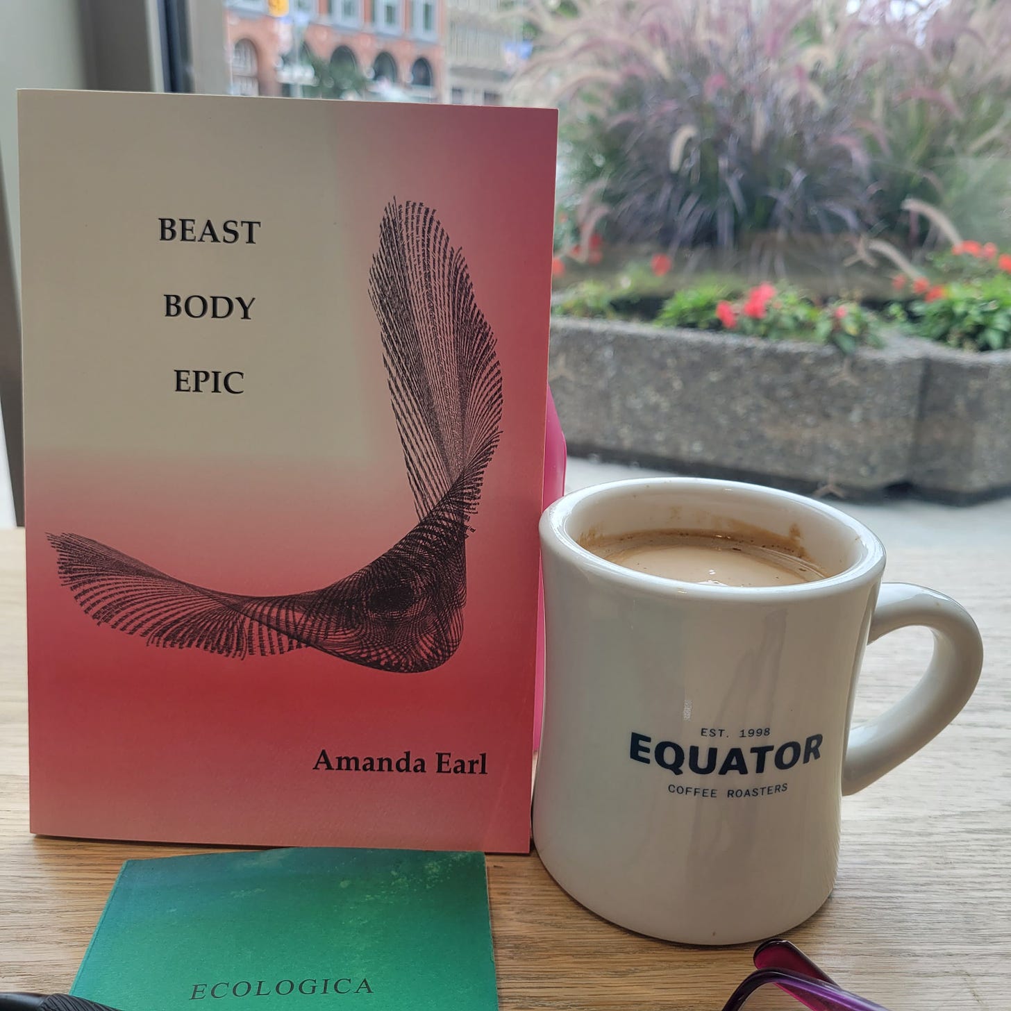 Book with red and white cover and an image of a visual poem. TEXT: BEAST/BODY/EPIC/Amaanda Earl beside an Equator coffee mug full of coffee and cream in front of a window that overlooks planters and a street with buildings across the street