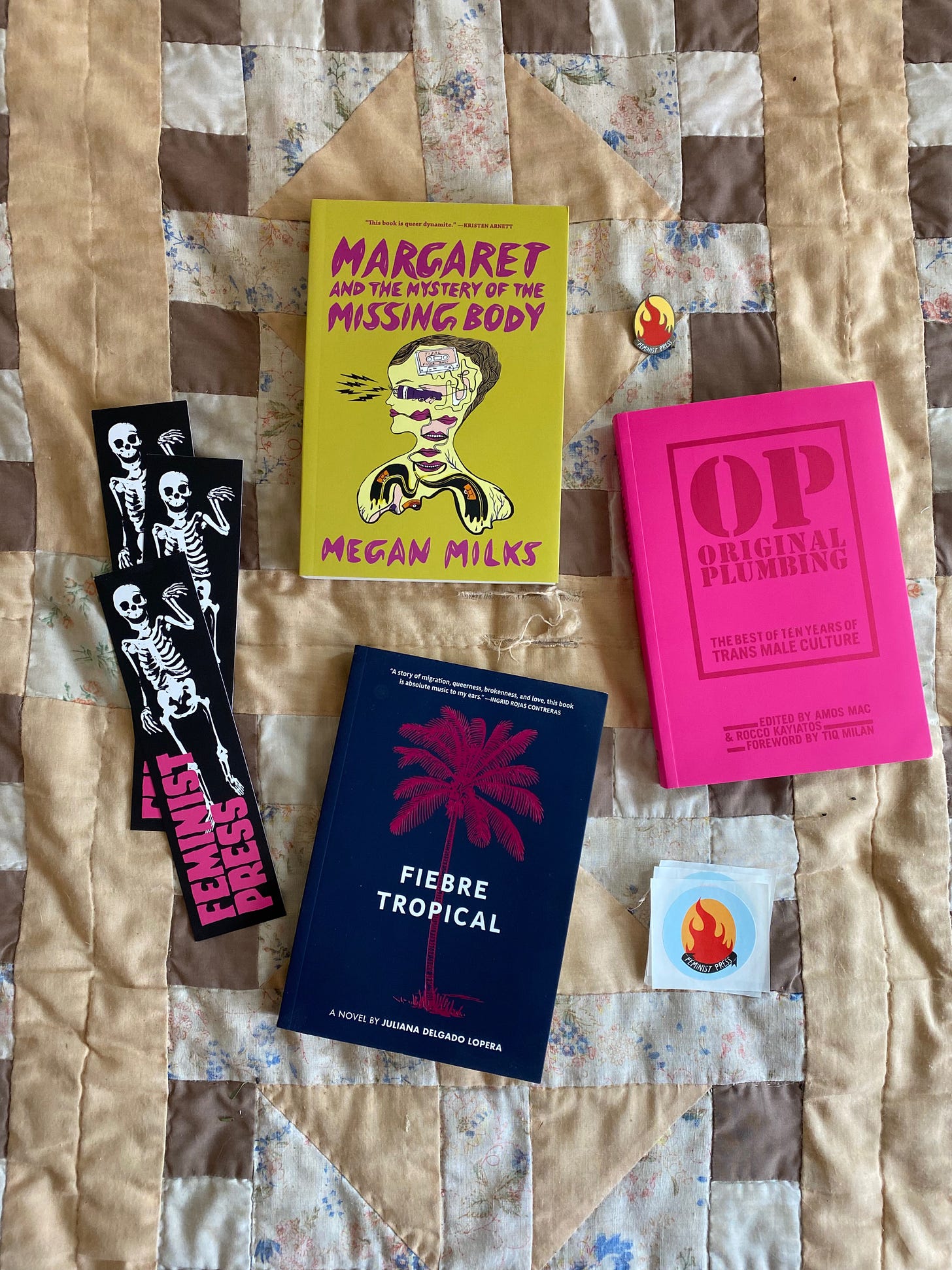 The three listed books arranged on a brown and tan quilt next to a small enamel pin with the Feminist Press logo and several black bookmarks with an illustration of a skeleton.