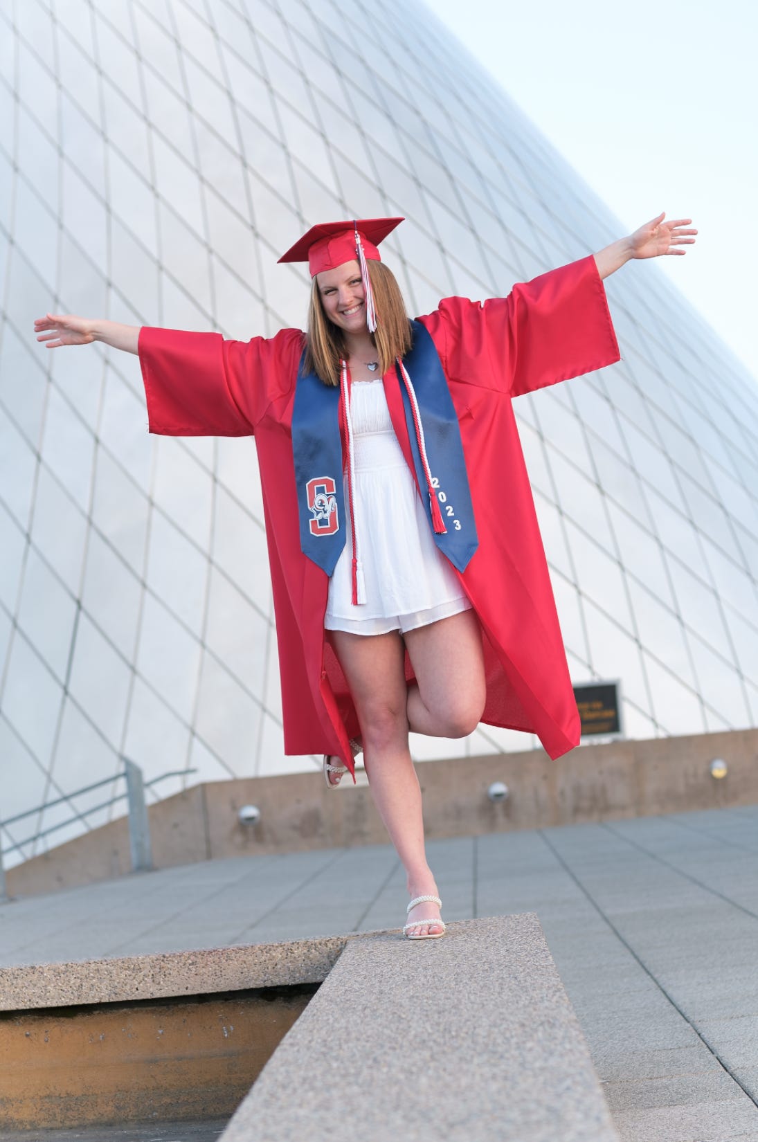 A young woman wearing a red cap and gown and a blue sash balances on one leg in front of the metal cone sculpture at the Museum of Glass