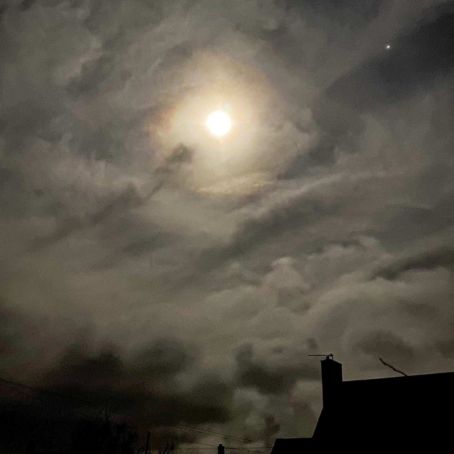 An image of the moon, glowing through clouds with a rooftop silhouetted below.
