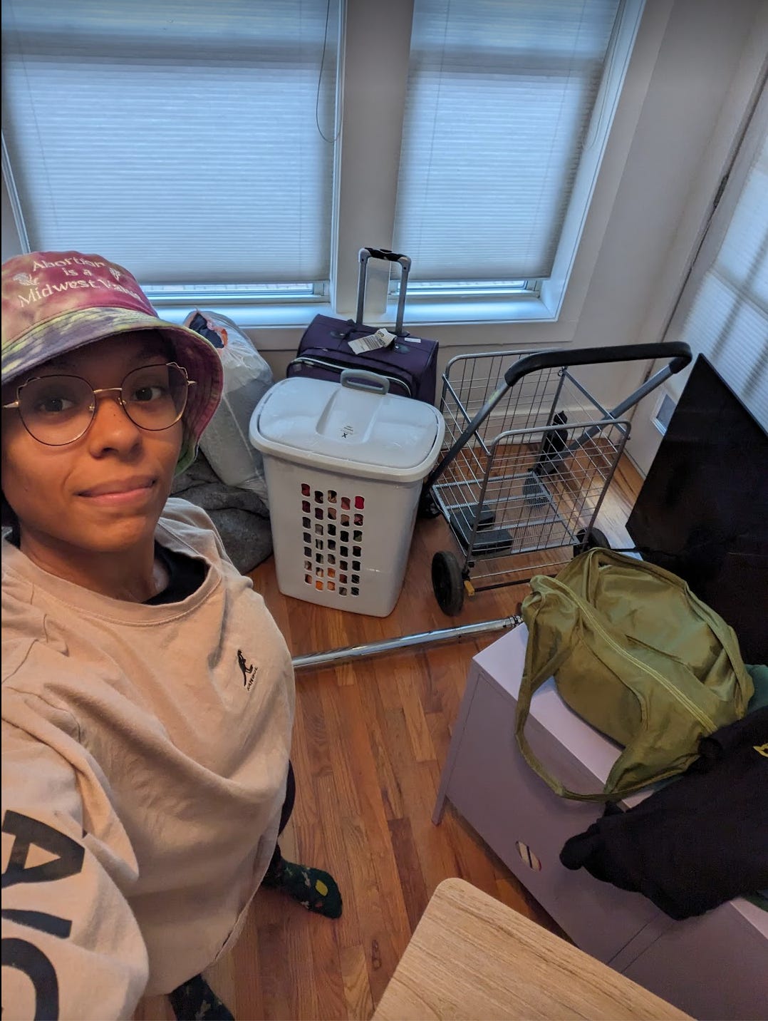 Nathalie takes a selfie of her new messy living room that thas bags, a laundry basket, cart and suitcases spread around. She is wearing a tie dyed bucket hat that says "Abortion is a Midwest Value"