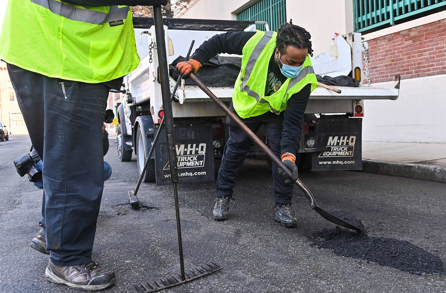 A public works team member wearing a neon safety vest uses a shovel to pile hot asphalt into the pothole to get ready for filling. Behind him, the white pick up truck has a heap of asphalt on the truck bed, covered by a tarp