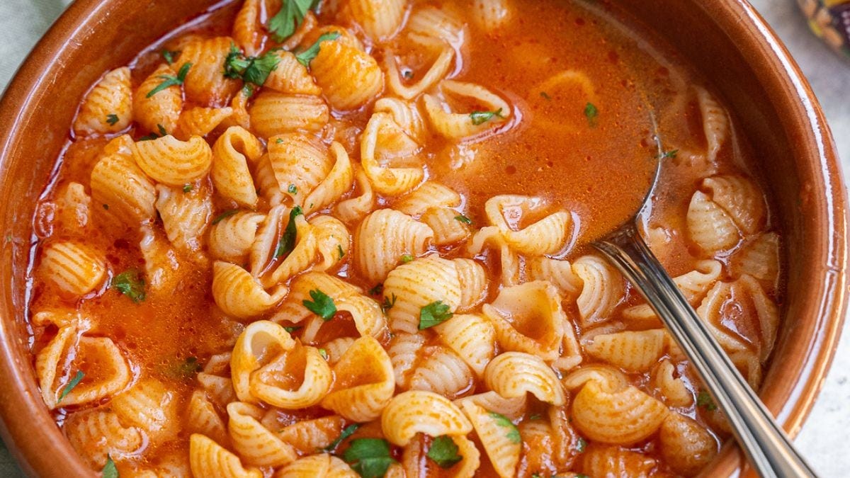 A bowl of tomato-based pasta soup with shell noodles, garnished with fresh herbs, photographed from above.