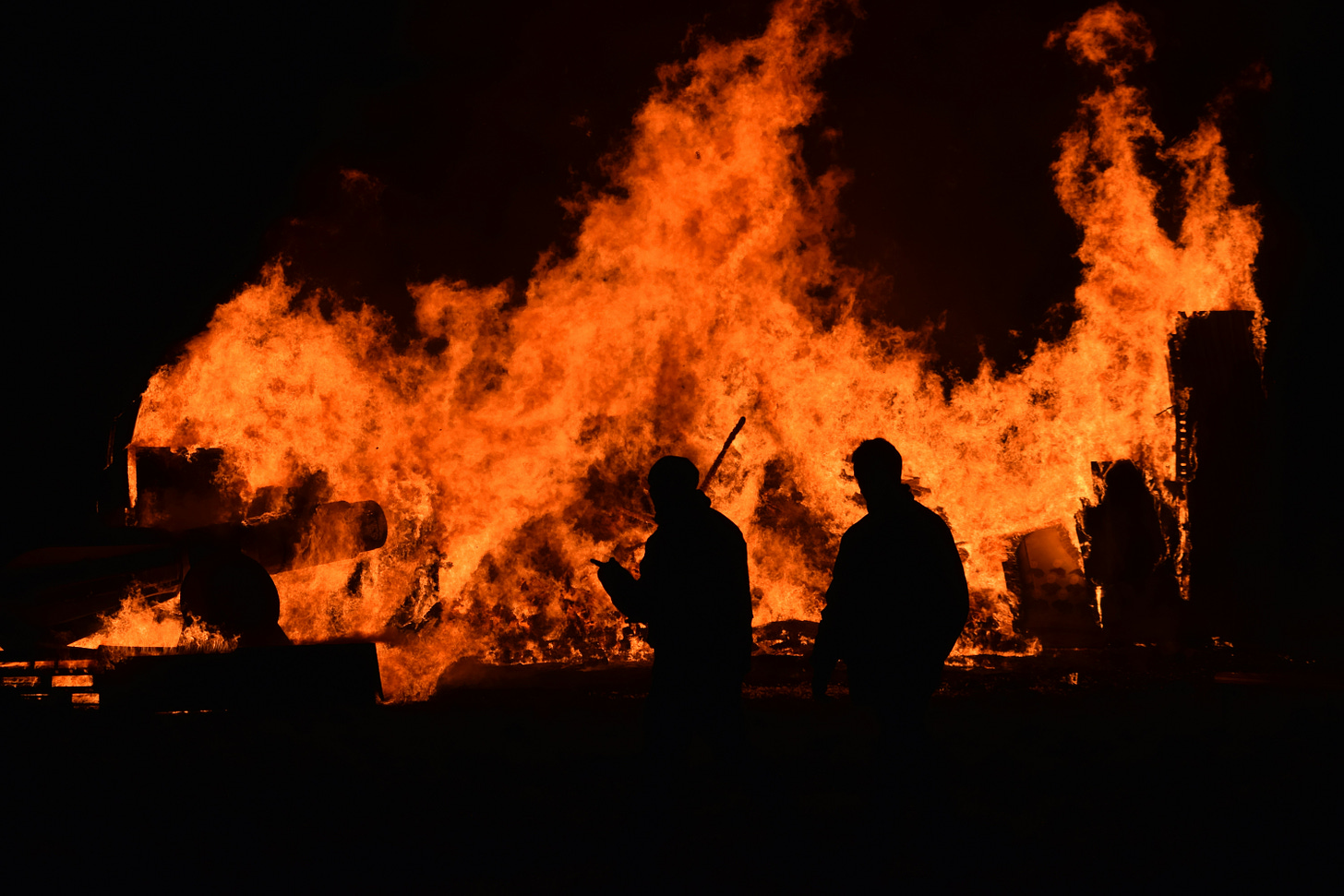 People walk in front of a large fire, in the dark.