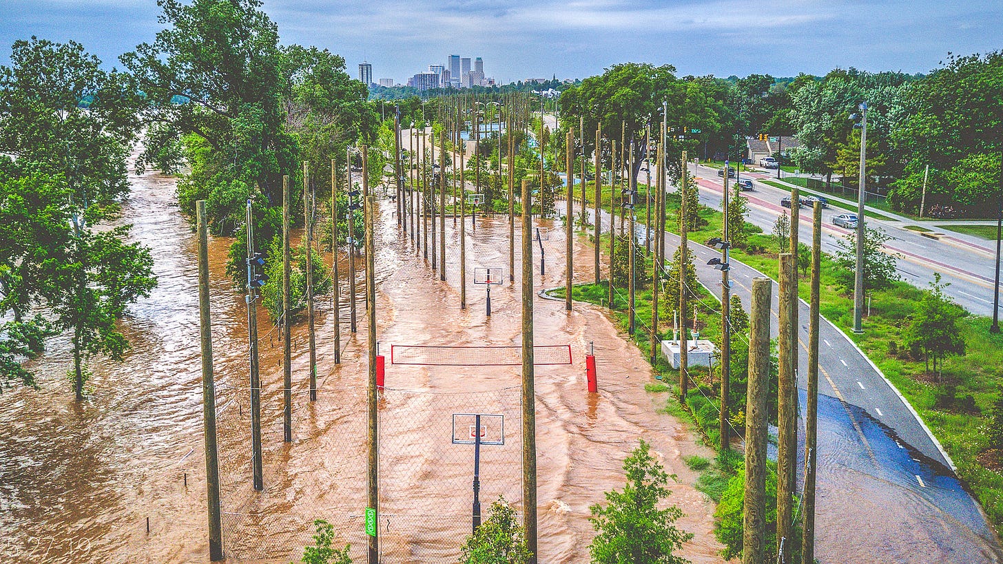 Mud-orange floodwaters lap across the sports courts at Gathering Place beneath numerous wood-clad light poles, bright green vegetation and a red volleyball net. The water spills across a portion of the paved trail as cars pass on Riverside Drive. 