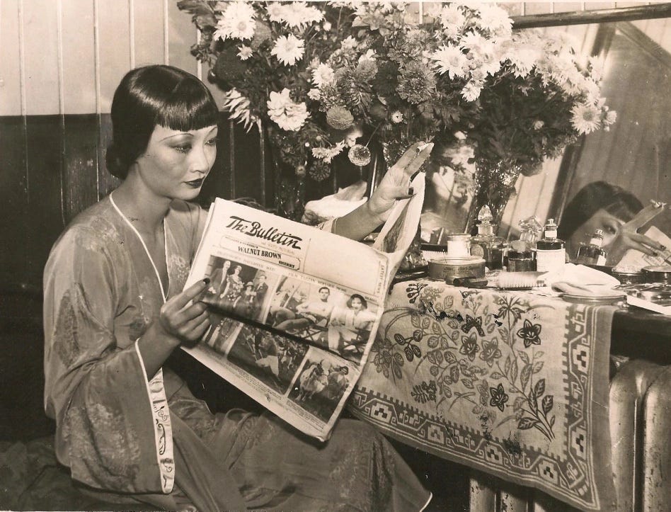 Anna May Wong in a silk robe sits at her backstage dressing table strewn with bouquets and beauty products, she reads The Bulletin newspaper held in her hands