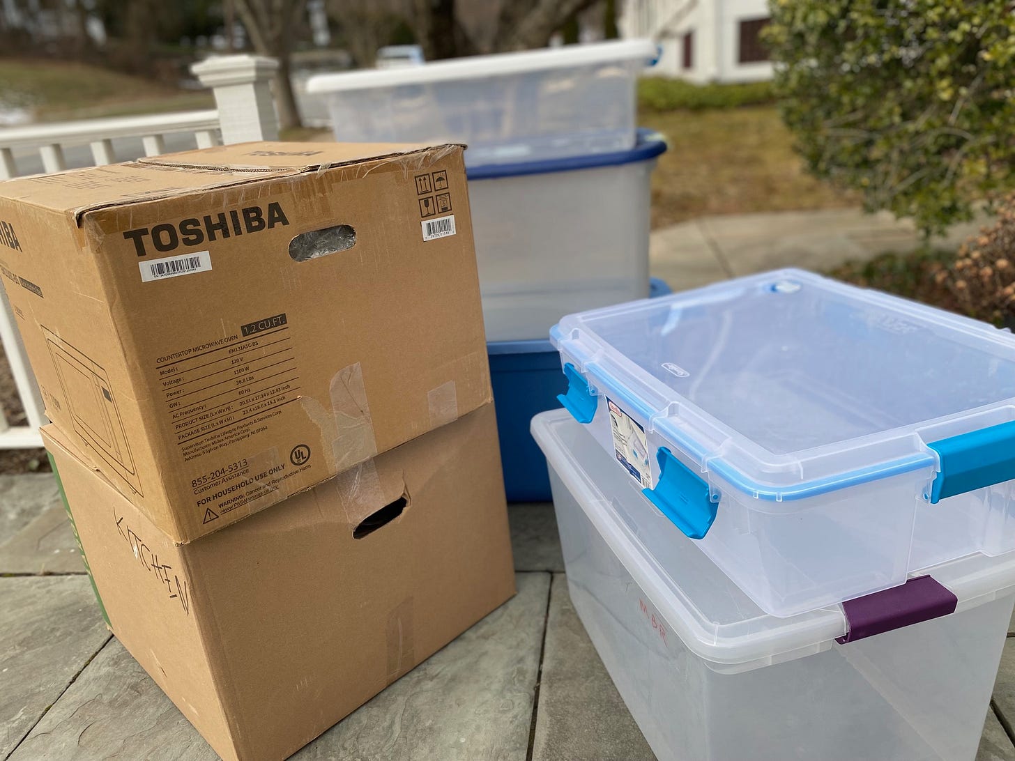 Two stacked brown boxes and five clear and empty plastic bins on slate sidewalk.