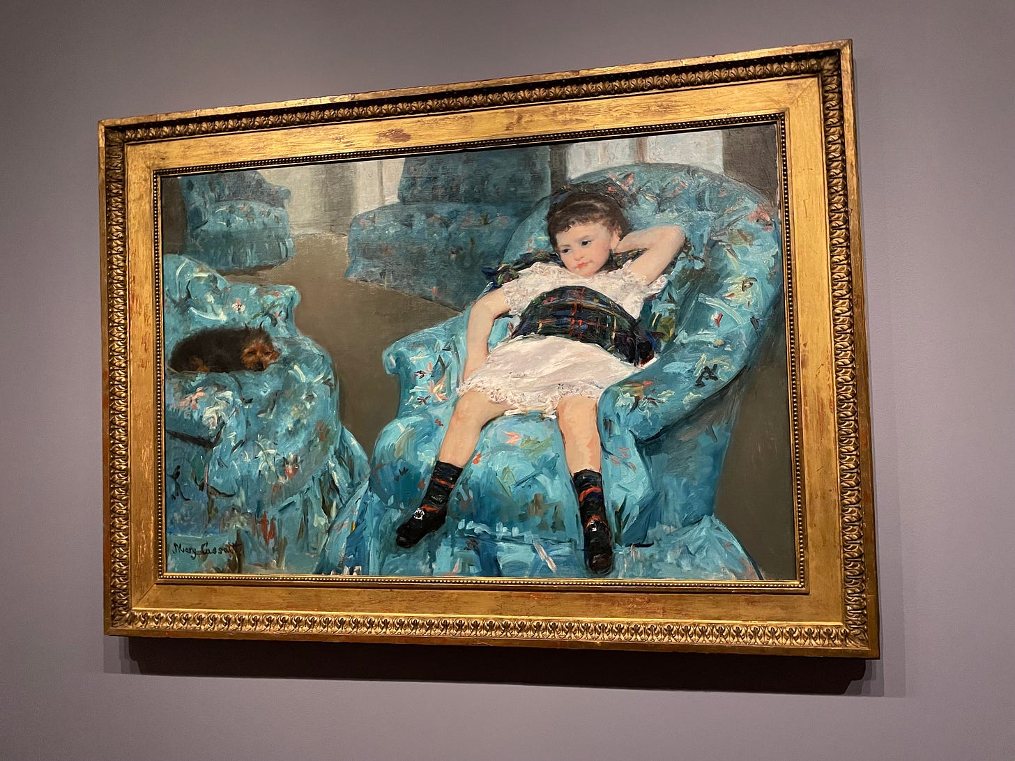 Photograph of Mary Cassatt's painting "Little Girl in a Blue Armchair" depicting a young girl slouching in a blue patterned armchair with an exhausted expression. She is wearing a plaid dress and plaid socks. She has brown hair. A brown dog is resting on an adjacent armchair. 