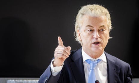 Photograph of Geert Wilders standing against a black background gesticulating and pointing with one hand as he speaks. He is wearing a dark blue suit jacket, white shirt and pale blue tie, and has swept-back blond hair
