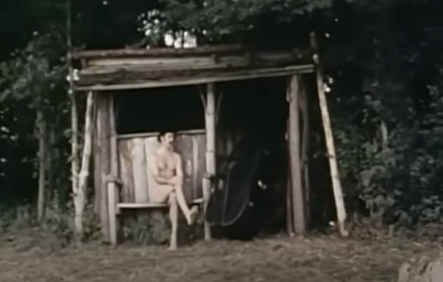 A man (John Cleese) sits nude on a wood bench before an old wooden shack. Still frame from Romance with a Double Bass (1974).